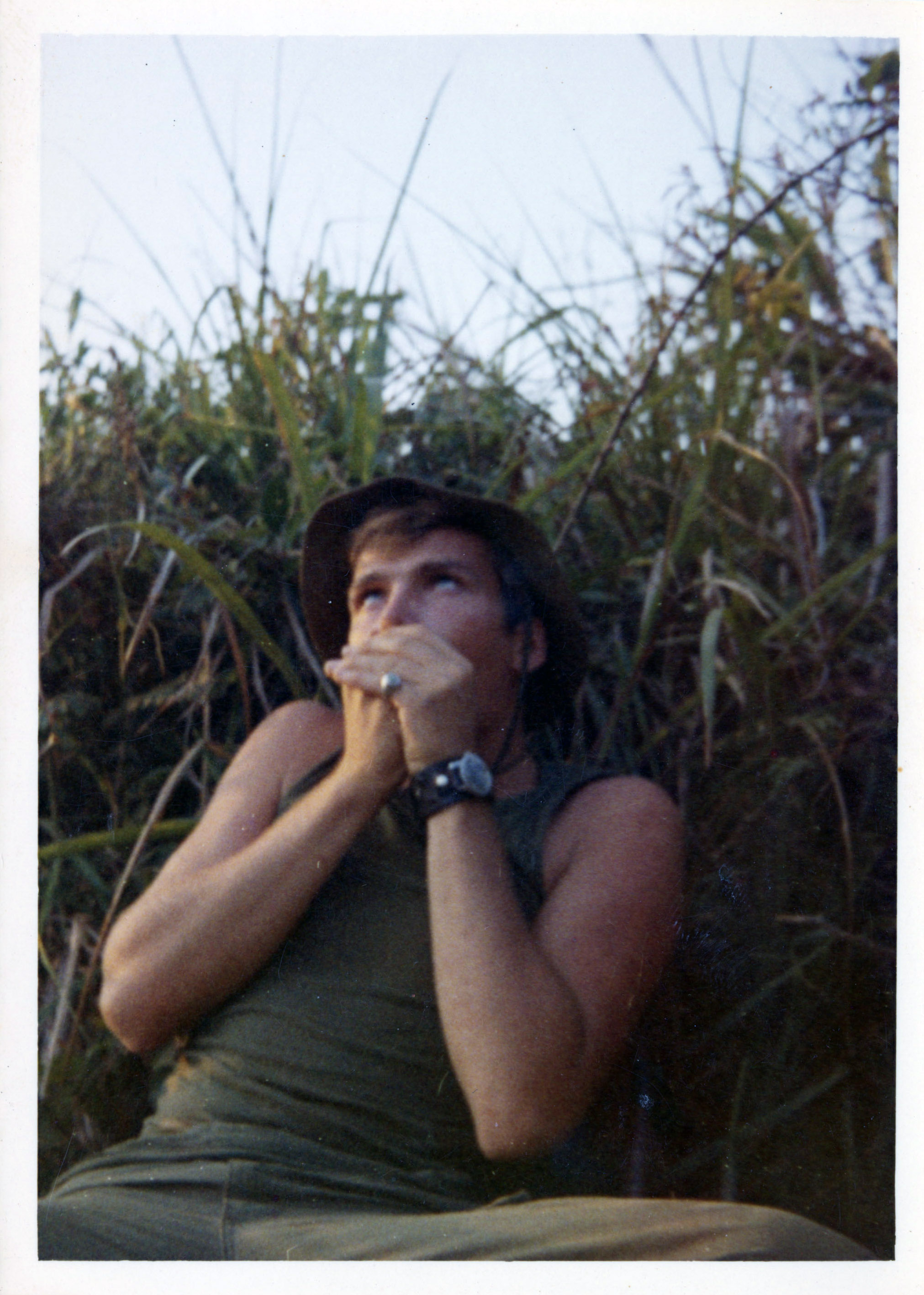 Terry posing for a camera while holding his hands over his mouth, potentially making some kind of a bird call, while sitting in tall grass.