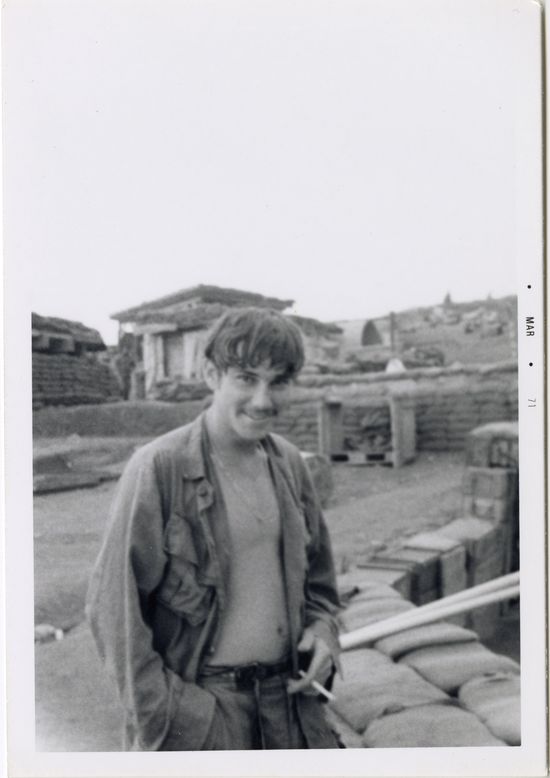 A soldier is posing at base camp with an open shirt and cigarette in his hand.