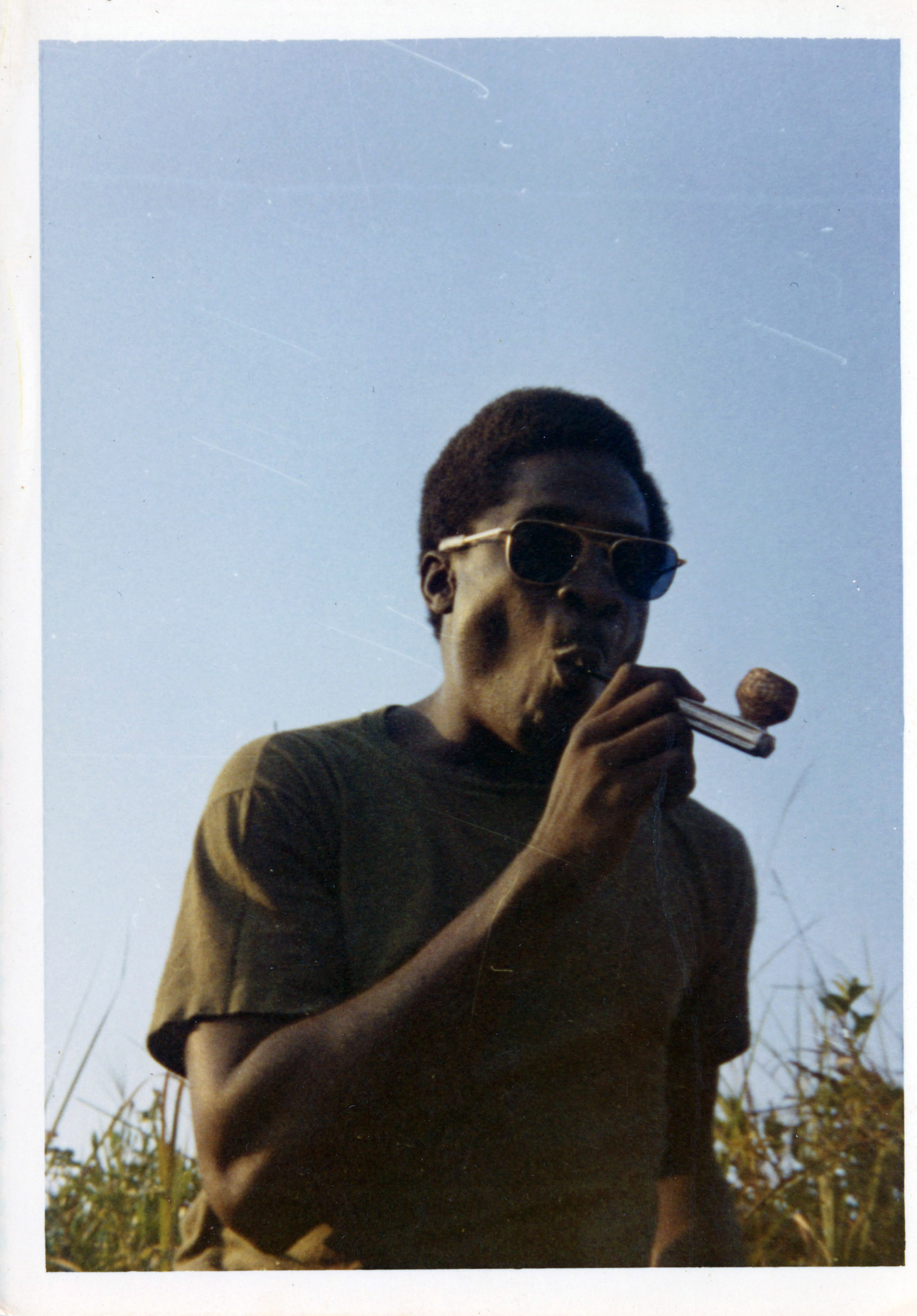 A soldier in the Vietnam war is smoking from a pipe while sitting down.