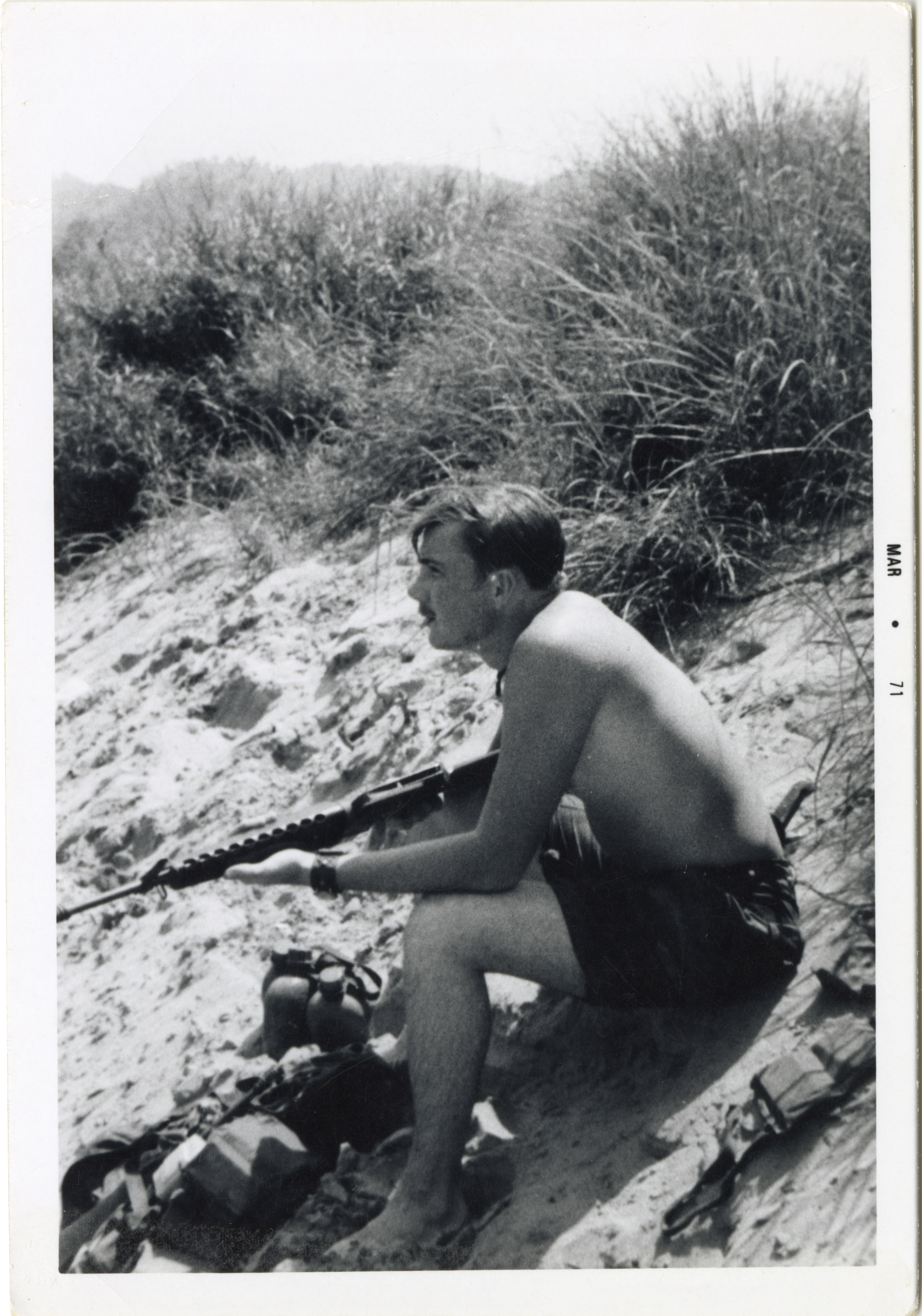 A profile view of Terry holding a semi automatic rifle, sitting on a hillside and staring into the distance.