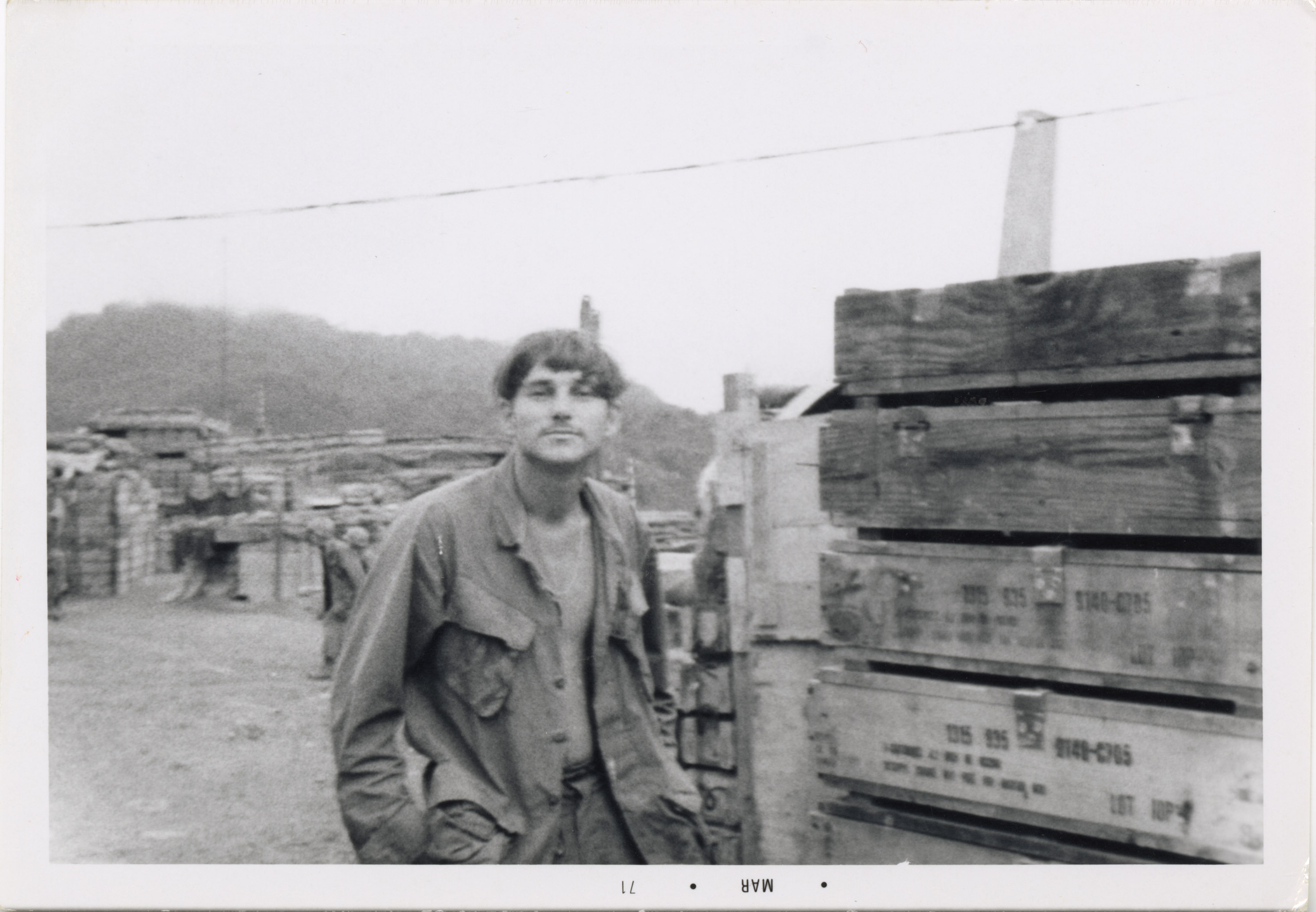 A soldier poses for the camera with an open shirt and is sitting at base camp.