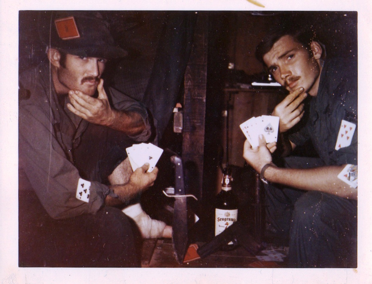 Terry and an unknown fellow soldier are playing cards, sharing their hands toward the camera, while striking a pose by holding their chins.