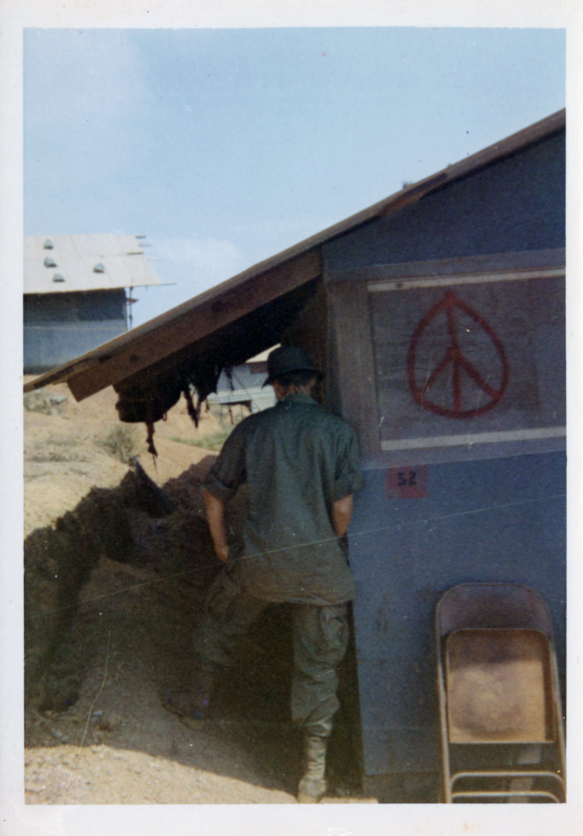 Soldier facing away from camera, standing next to a hooch with a peace symbol spray painted in red on the side of it.