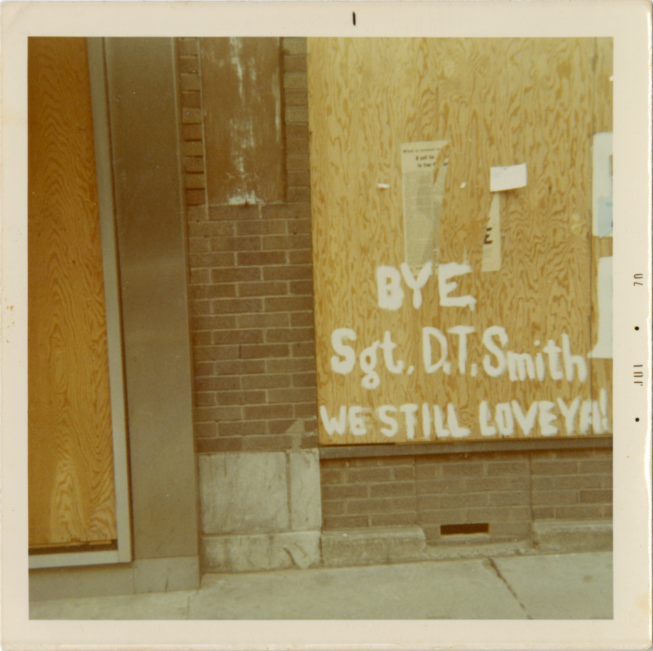 The words "BYE, Sgt. D.T. Smith WE STILL LOVE YA!" are painted in white on the side of a building.