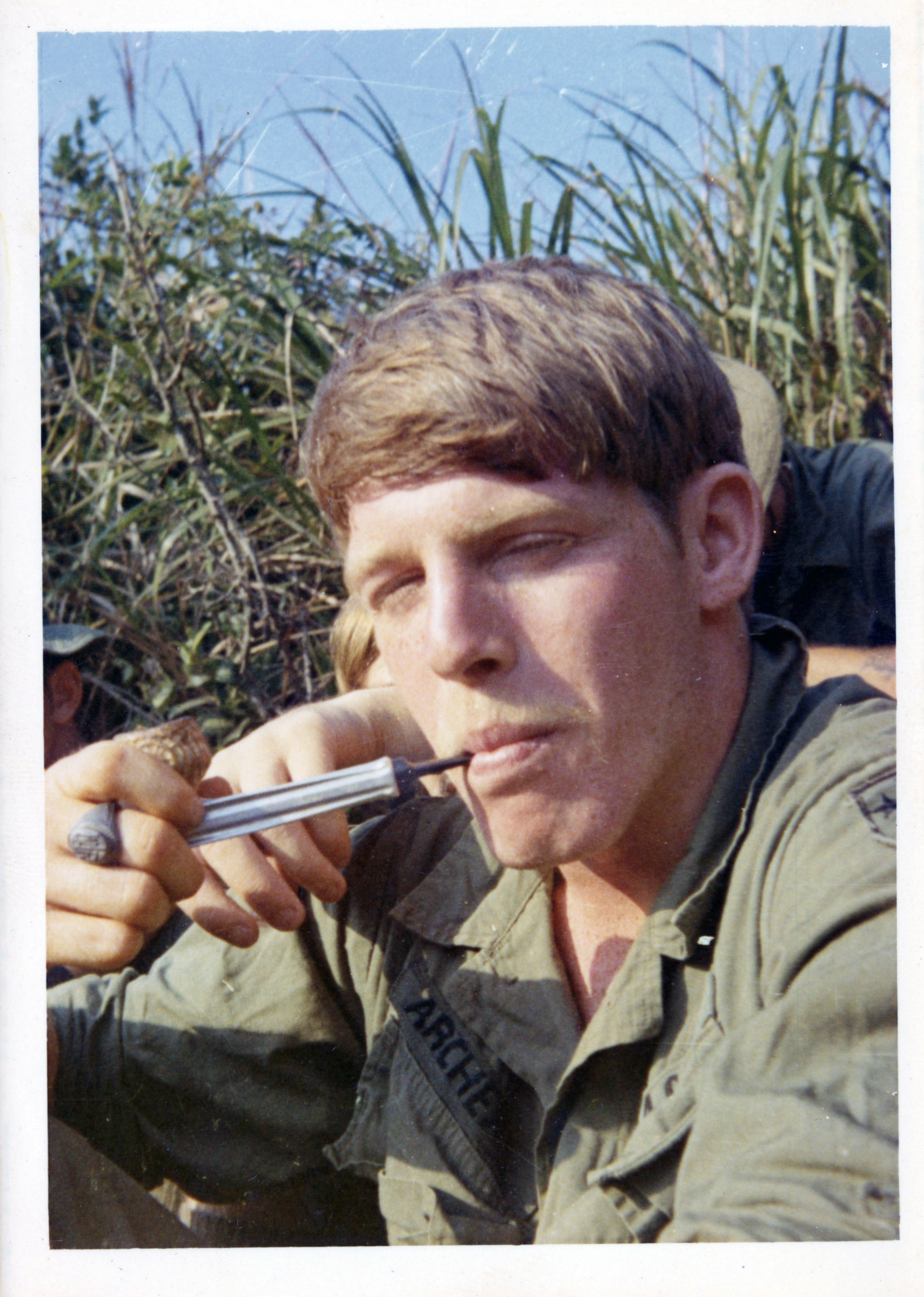 A soldier smokes from a pipe while staring directly at the camera, squinting his eyes.